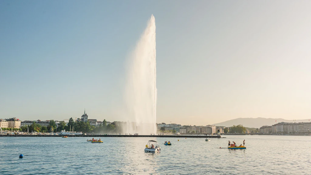 Vista de um lago com barcos e o jato de água no centro, edifícios na margem e céu limpo.