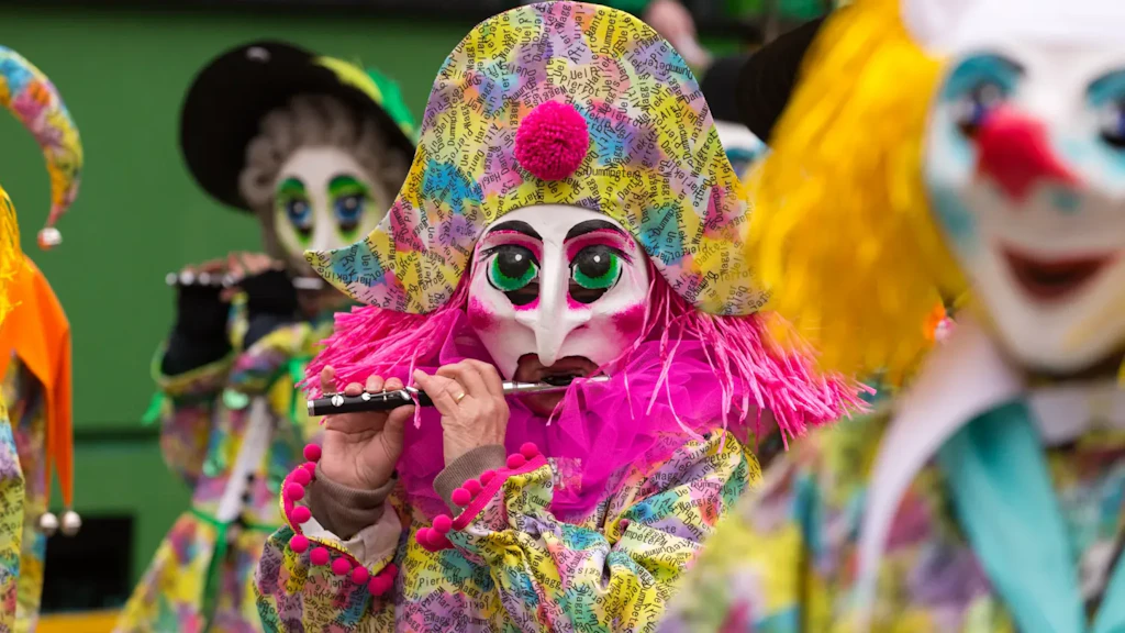 Pessoa mascarada com fantasia colorida e chapéu grande tocando flauta, com outras figuras fantasiadas ao fundo.
