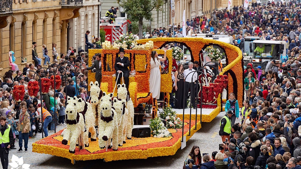 Um carro alegórico decorado festivamente com ornamentos florais e vacas artificiais desfilapor uma multidão densa de pessoas.