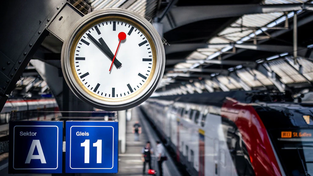 Relógio da SBB com mostrador branco, ponteiros pretos das horas e dos minutos e ponteiro vermelho dos segundos.
